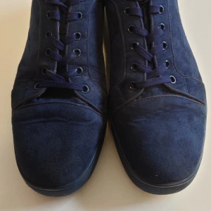 Mörkblå sneakers från Christian Louboutin - Säljer ett par mörkblå sneakers från Christian Louboutin med klassisk röd sula och snörning. Skorna är i mocka med rund tå och har en beige insida i skinn. Perfekta för dig som vill sticka ut med exklusiva detaljer.