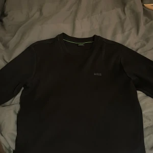 Svart sweatshirt från BOSS - Säljer en stilren svart sweatshirt från BOSS med diskret logga på bröstet. Tröjan har rund halsringning och långa ärmar. Perfekt för en clean och enkel look.