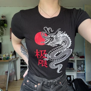 Svart t-shirt med draktryck  - Svart ribbad t-shirt drakmotiv. T-shirten har korta ärmar och rund halsringning. Perfekt statement-plagg för en cool look.