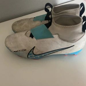 Nike Air Zoom fotbollsskor i vitt och blått - Säljer ett par Nike Air Zoom fotbollsskor i vitt med blå detaljer och svart swoosh. Skorna har en elastisk mudd, bred kardborre över vristen och mönstrad sula i blått och rött. Perfekta för dig som vill sticka ut på planen!