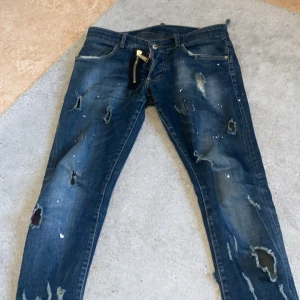 Blå slitna jeans från Dsquared2 med dragkedjedetalj - Säljer ett par mörkblåa blå jeans från Dsquared2 med slitningar, hål och färgstänk för en riktigt edgy look. Byxorna har en unik dragkedjedetalj framtill och klassisk femficksdesign. Perfekta för dig som gillar streetstyle och vill sticka ut. Bixorna är såklart äkta.  Sitter bra på mig som är 1,69 cm lång ish. Jag säljer det därför att jag inte har det till användningen längre det går såklart att byta om du har nått snyggt som passar mig  