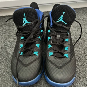 Svarta och blå Jordan sneakers - Säljer ett par svarta Jordan sneakers med blå detaljer och blå sula. Skorna har det klassiska Jumpman-loggan i turkos på plös, häl och sula. Ovandelen är i syntetmaterial med snörning och sportig design. Perfekta för dig som gillar streetwear och basketstil.