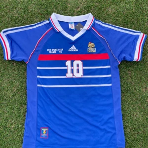 Frankrike 1998 World Cup Hemma Kit - Klassisk blå Frankrike fotbollströja från VM 1998 med Zidane och nummer 10 på ryggen. Adidas-logga, FFF-emblem och vita samt röda detaljer. Kortärmad med vita och röda ränder på ärmarna och flera horisontella ränder på framsidan.