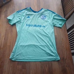 Ljusblå Schalke 04 fotbollströja från Adidas - Säljer en ljusblå Schalke 04 fotbollströja från Adidas med klubbmärke och sponsortryck på bröstet. Tröjan har korta ärmar och blå detaljer på axlarna. Perfekt för dig som gillar tysk fotboll!