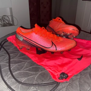 Röda Nike Mercurial fotbollsskor - Säljer ett par röda Nike Mercurial fotbollsskor med svart swoosh och detaljer. Säljer det här gamla paret för att den sitter och ligger på hyllan bara. Skon har några defekter på hällen och lite smutsig sula men annars är den perfekt. Pris kan diskuteras!