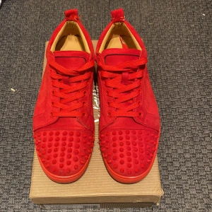 Röda sneakers med nitar från Christian Louboutin - Säljer ett par helröda sneakers från Christian Louboutin med mockaöverdel och matchande röda nitar på tån. Skorna har snörning och den klassiska röda sulan med logotyp. En nit har lossnat på högra skon längst fram i mitten. Priset kan diskuteras vid snabbare affär