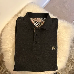 Mörkgrå pikétröja från Burberry - !! PASSAR INTE XL, PASSAR S / M har själv storlek S !! Säljer en mörkgrå pikétröja från Burberry med klassisk krage och knäppning framtill. Tröjan har Burberrys rutiga mönster på insidan av kragen och en broderad logga på bröstet. Perfekt för en stilren och tidlös look.