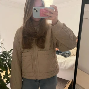 Beige quiltad jacka - Säljer en beige quiltad jacka med dragkedja och ribbade muddar. Jackan har två stora fickor framtill och en elegant passform. Perfekt för dig som gillar en stilren och enkel look.