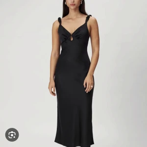 Svart Nimes slip dress från Adoore - Säljer en svart långklänning från Adoore med smickrande passform och twistad detalj vid axelbanden. Klänningen har en liten öppning framtill och är ärmlös med figurnära siluett. Perfekt för speciella tillfällen.