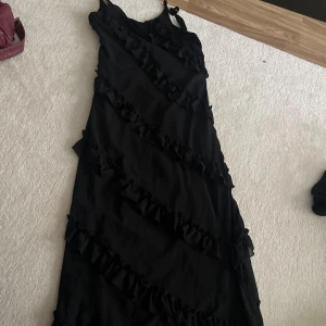 Svart långklänning med volanger - Säljer en svart långklänning med smala axelband och dekorativa volanger som löper diagonalt över hela klänningen. Perfekt för dig som vill ha en elegant och stilren look med lite extra detaljer.