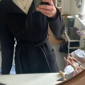 Svart kappa med bälte - Snygg svart kappa med bälte som ger en elegant look. Perfekt för kyligare dagar med sin långa design och klassiska stil. Passar bra till både jeans och mer formella outfits.