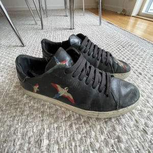 Svarta sneakers med broderade fåglar från Axel Arigato - Säljer dessa skor från Axel Arigato, mörkblå/svart med röda och blåa broderade fåglar. Skorna är använda en del och på bilden ser man vilka slitningar som finns. Detta är dock inget som syns när man har på sig skorna. Nypriset var 2300 kr när jag köpte dem så säljer för ett bra pris!