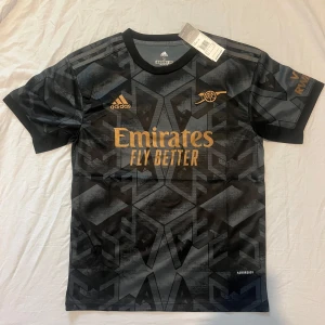 Svart och grå fotbollströja från Adidas - Snygg svart och grå fotbollströja från Adidas med ett geometriskt mönster. Tröjan har korta ärmar och guldiga detaljer med texten 'Emirates Fly Better' och 'Visit Rwanda'. Perfekt för fotbollsfans som vill ha en stilren look.