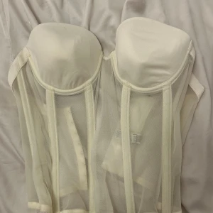 Vit korsett med meshdetaljer - Elegant vit korsett med vadderade kupor och transparenta meshpaneler. Perfekt för att skapa en stilren och sofistikerad look. Den har en klassisk design med en modern twist.