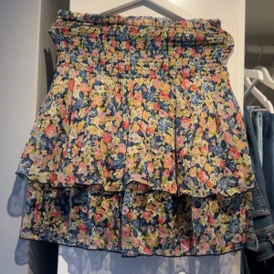 Blommig kortkjol med volang - Charmig blommig kortkjol med volangdetaljer. Kjolen har en elastisk midja för bekväm passform och ett färgglatt blommönster.