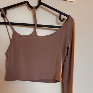 Brungrå/beige one-shoulder cropped topp - Brungrå one-shoulder cropped topp med lång ärm. Perfekt för festliga tillfällen. Tröjan har en unik stil och är i väldigt bra skick! Passar extremt bra med både kjolar och jeans.