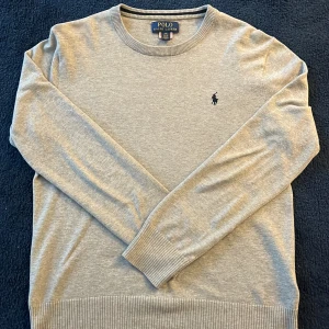 Grå tröja från Polo Ralph Lauren - Säljer en stilren grå tröja från Polo Ralph Lauren. Tröjan har en klassisk rund halsringning och är dekorerad med den ikoniska logotypen på bröstet. Den är tillverkad i ett mjukt material med ribbade muddar vid ärmslut och nederkant. Ny pris för denna tröja är ca 1450kr