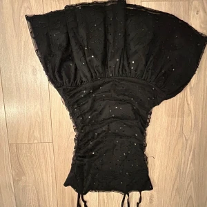 Svart klänning från Gina Tricot - Snygg svart klänning från Gina Tricot med paljettdetaljer och smala axelband. Klänningen har en åtsittande passform och är perfekt för festliga tillfällen. Den har en elegant rynkad design som ger en fin siluett. Pris kan diskuteras