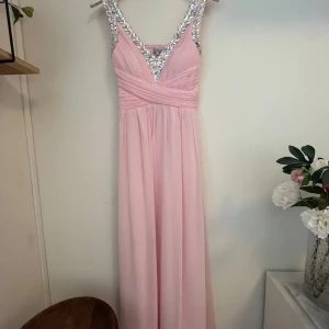 Rosa långklänning från Scala - Elegant rosa långklänning från Scala med v-ringad design och glittriga detaljer på axelbanden. Klänningen har en omlottstil i midjan och är perfekt för speciella tillfällen. Inkluderar en matchande sjal för extra stil.ä. Använd en gång! En pärla är lös (se bild) men går att limma dit, därav säljs den billigt. Köpt för 1500kr.