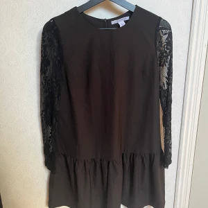 Svart klänning med spetsärmar - Elegant svart klänning med långa spetsärmar och en volangdetalj vid fållen. Perfekt för en stilren look med en touch av romantik. Klänningen har en rund hals och en dragkedja i ryggen.