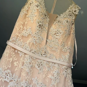 Champagnefärgad spetsklänning med pärlor - Elegant champagnefärgad klänning med vacker spets och pärldetaljer. Klänningen har en v-ringad design och ett smalt bälte i midjan. Perfekt för speciella tillfällen.