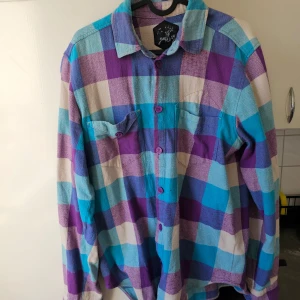 Rutig skjorta från Public Gallery - Säljer en färgglad rutig skjorta från Public Gallery i storlek L. Skjortan har en mix av lila, blå och beige rutor och är tillverkad i 100% bomull. Den har långa ärmar och knappar framtill samt på ärmsluten. Perfekt för en avslappnad stil!