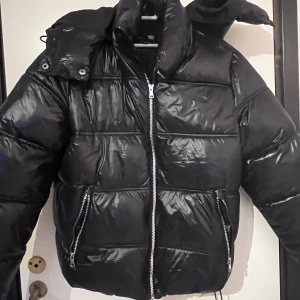 Svart pufferjacka med dragkedja - Säljer en svart pufferjacka med glansig yta och dragkedja framtill. Jackan har en hög krage och avtagbar huva. Perfekt för kyliga dagar! Storlek xs bara provad aldrig använd! Pris kan diskuteras 