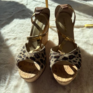 Leopardmönstrade kilklackar med gulddetaljer - Snygga kilklackar med leopardmönster och guldfärgade remmar. Skorna har en öppen tå och en hög korkplattform som ger extra höjd. Perfekta för att sätta en trendig touch på din outfit.