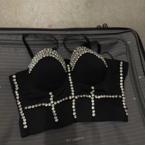 Svart korsettopp med strassdetaljer - Snygg svart korsettopp dekorerad med glittrande strassdetaljer. Toppen har justerbara axelband och en snörning bak för perfekt passform. Perfekt för en kväll ute eller en speciell tillställning.