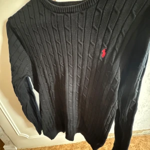 Svart ribbad tröja från Polo Ralph Lauren - Säljer en stilren svart ribbad tröja från Polo Ralph Lauren med det klassiska röda logotypen på bröstet. Tröjan har långa ärmar och en rund halsringning, perfekt för en elegant och avslappnad look. L/XL