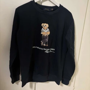 Svart tröja med Polo Bear från Ralph Lauren - Säljer en svart långärmad tröja från Ralph Lauren med det ikoniska Polo Bear-motivet på framsidan. Tröjan har rund hals och ribbade muddar vid ärmslut och nederkant. Perfekt för en stilren och avslappnad look.