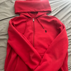 Röd hoodie från Ralph Lauren - Säljer en snygg röd hoodie från Ralph Lauren med dragkedja och klassisk logga på bröstet. Perfekt för en avslappnad stil. Den har en bekväm passform och en praktisk huva med snörning.