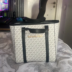 Vit och svart axelväska från Michael Kors - Snygg axelväska från Michael Kors i vitt och svart med det klassiska MK-mönstret. Väskan har svarta handtag och detaljer i guld. Insidan är rymlig med flera fack och dragkedja för säker förvaring. Perfekt för att bära med sig det nödvändigaste med stil.