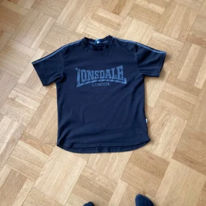 Svart t-shirt från Lonsdale - Säljer en svart t-shirt från Lonsdale med tryck på framsidan. T-shirten har korta ärmar och en klassisk rund halsringning. Perfekt för en avslappnad stil.