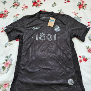 Svart fotbollströja från Nike AIK - Säljer en svart fotbollströja från Nike med tryck '1891' på framsidan. Tröjan är kortärmad och tillverkad i 100% polyester med Dri-FIT teknologi för optimal komfort. Perfekt för fotbollsfans som vill ha en stilren look på planen.