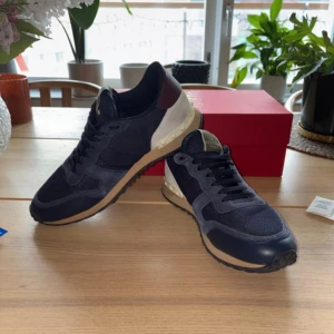 Valentino Rockrunner sneakers i blått och vinrött - Snygga Valentino Rockrunner sneakers i en stilren kombination av blått och vinrött. Skorna har en klassisk snörning och är tillverkade i en mix av mocka och syntetmaterial. De har en unik detalj med nitar på sidan och en bekväm sula i beige.