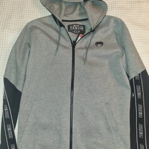Venum zip hoodie - Snygg grå hoodie från Venum med svarta detaljer och dragkedja. Ärmarna har Venum-logga längs sidorna och huvan är justerbar med snören. 