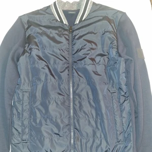 Hugo Boss cardigan - Mörkblå jacka från Boss med dragkedja och ribbad krage. Jackan har långa ärmar och en quiltad design framtill. 