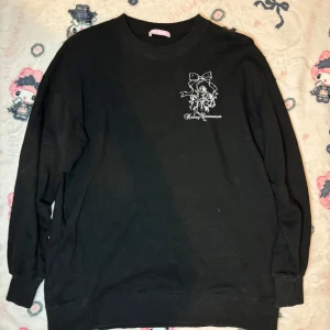 Svart tröja från Honey Cinnamon - Snygg svart tröja från Honey Cinnamon med tryck av rosett och änglar både fram och bak. Textdetaljer ger en unik touch. Perfekt för en stilren och casual look. Oversized 