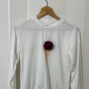 Vit långärmad tröja med glassmotiv - Säljer en vit långärmad tröja med ett unikt glassmotiv på framsidan. Motivet består av en röd boll som ser ut som en glasskula på en guldfärgad strut. Perfekt för en lekfull stil!