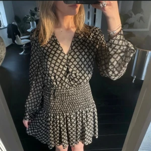 Mönstrad klänning med långa ärmar - Snygg klänning med ett geometriskt mönster i svart och beige. Klänningen har långa ärmar och en smickrande v-ringad hals. Den har en smockad midja som ger en fin passform och en lätt utställd kjol.