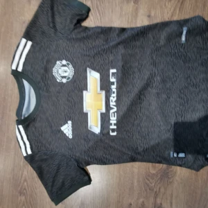 Svart Manchester United fotbollströja från Adidas - Säljer en svart Manchester United fotbollströja från Adidas med korta ärmar och vita detaljer. Tröjan har klubbens emblem på bröstet och ett mönster över hela tyget. Perfekt för fans som vill visa sitt stöd.