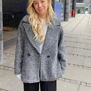 Kappa från design by si - Säljer en stilren grå och svart rutig kappa med stora slag och knappar framtill. Perfekt nu till våren då den är rätt tunn.
