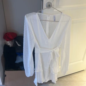 Vit klänning från Amisu - Säljer en elegant vit klänning från Amisu med v-ringning och knytband i midjan. Klänningen har långa ärmar och ett subtilt prickigt mönster. Perfekt för sommarens festligheter!