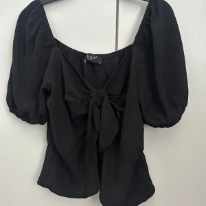Svart blus med puffärmar - Snygg svart blus från sisters point med puffiga kortärmar och en knytning framtill. En volangdetalj vid nederkanten. Perfekt för en stilren look. 