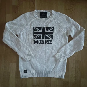 Vit stickad tröja från Morris - Säljer en stilren vit stickad tröja från Morris med ett stort svart och blått Union Jack-motiv på bröstet. Tröjan har långa ärmar och ribbade muddar vid ärmslut och nederkant. Perfekt för en avslappnad och trendig look.