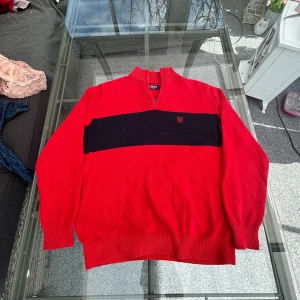 Röd tröja med dragkedja från Chaps - Säljer en snygg röd tröja från Chaps med en svart horisontell rand över bröstet och ett broderat emblem. Tröjan har en dragkedja vid halsen och ribbade muddar vid ärmslut och nederkant. Perfekt för en stilren look!