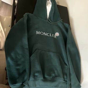 Grön hoodie från Moncler - Säljer en snygg grön hoodie från Moncler med vit logotyp på bröstet. Den har en praktisk magficka och en justerbar huva med vitt foder. Perfekt för en avslappnad stil.
