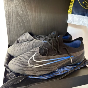 Nike phantom gx elite fotbollskor - Snygga fotbollsskor från Nike i svart med blå och vita detaljer. Skorna har en modern design med snörning och en strömlinjeformad siluett. Perfekta för fotbollsplanen med en stilren look.