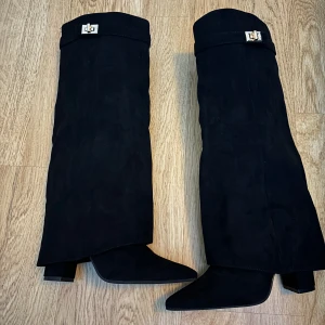 Svarta stövlar med dekorativt guld spänne - Snygga helt oanvända svarta stövlar i mocka med dekorativt spänne upptill. De har en spetsig tå och dragkedja på baksidan för enkel på- och avtagning. Perfekta för en stilren look. Urspruckspris 1 599kr